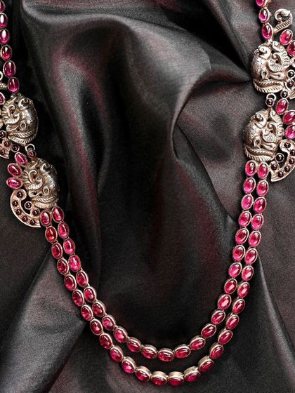 This handcrafted silver necklace features two strands of red stones, likely rubies, accented with peacock-patterned pendants. It is part of an exquisite set that includes matching earrings and a bracelet.