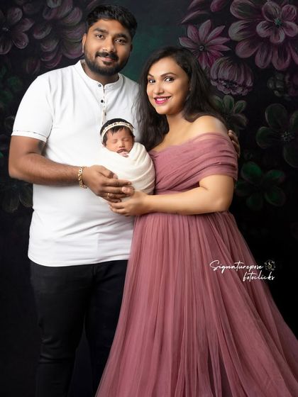 A stunning portrait of a new family. The mother, dressed in a flowing gown, holds her newborn while her husband stands proudly by her side.
