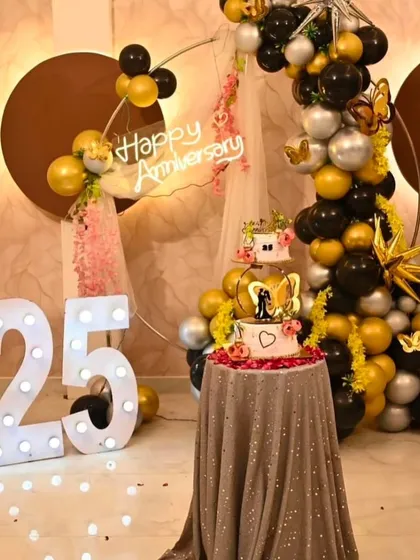 A grand 25th anniversary celebration. The decor includes a balloon arch, a "Happy Anniversary" neon sign, a tiered cake, and a large light-up number '25'.