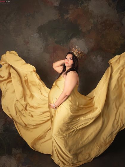 A powerful and artistic maternity portrait. The mom-to-be poses with a flowing golden gown that seems to fly around her, creating a stunning visual against a painted backdrop.