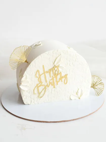 A modern and elegant half-birthday cake. The unique semi-circular shape is finished with a white textured frosting and accented with gold fans and a "Happy Birthday" topper.