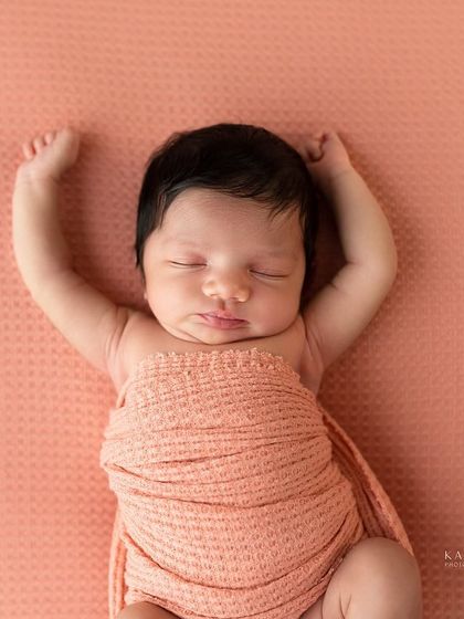 A one-week-old baby girl stretches in her sleep. The simple pink wrap and background keep the focus on this sweet, unposed moment.