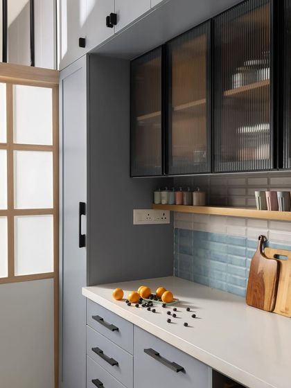 This compact kitchen blends pastel blue laminates with sleek black accents and warm wood finishes. A frosted glass sliding door separates the kitchen from the utility area, allowing natural light to flow through.