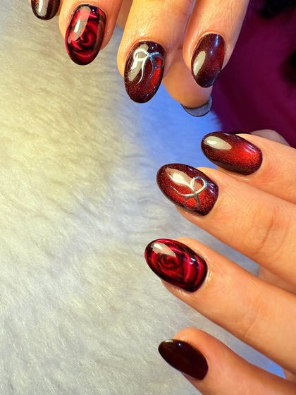 A dark and romantic nail set. This design features a deep red, almost black, cat-eye base with intricate rose and bow details, creating a moody and elegant look.