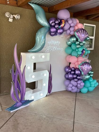 An outdoor mermaid-themed 5th birthday setup. A large marquee number "5" is the centerpiece, decorated with a mermaid tail balloon and an ocean-colored balloon garland.