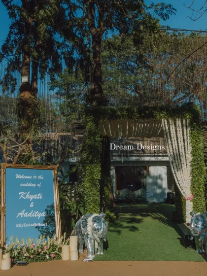 A daytime view of the grand wedding entrance. The welcome sign is nestled among flowers and candles, while decorative elephants stand guard at the leafy, flower-draped archway.