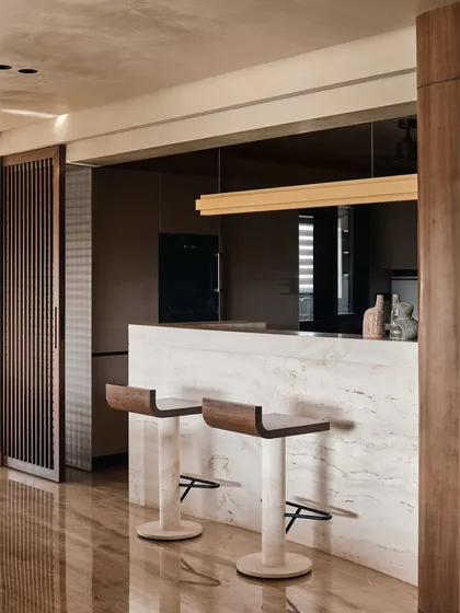 A minimalist bar area featuring a monolithic marble counter and sleek bar stools. A sliding wooden screen allows the space to be either open to the living area or concealed for a cleaner look.