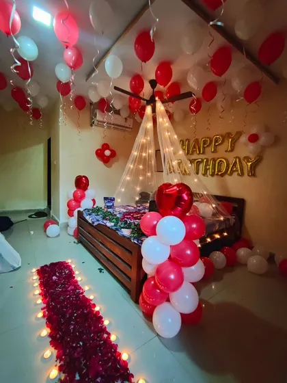 A complete romantic birthday surprise decoration for a bedroom. The setup includes a flower petal pathway with candles, a canopy over the bed with fairy lights, and red and white balloons everywhere.