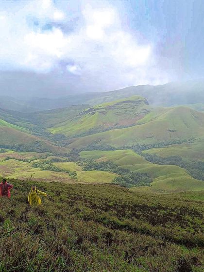 Two trekkers in colorful ponchos descending a trail, surrounded by the endless green valleys of Kudremukha.