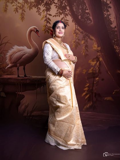 A graceful portrait of a mom-to-be in a traditional cream and gold saree, set against a painted swan backdrop for an artistic and cultural touch.