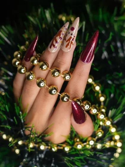A festive Christmas look featuring sculpted stiletto extensions. This set combines deep red glitter nails with nude accent nails, which are detailed with hand-painted reindeer art for a touch of holiday magic.