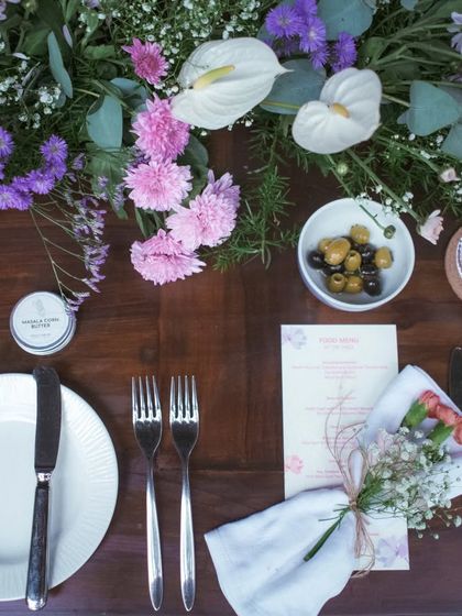 A stunning overhead shot of a tablescape adorned with a lush runner of purple and white flowers, with olives and personalized butter adding a thoughtful touch.