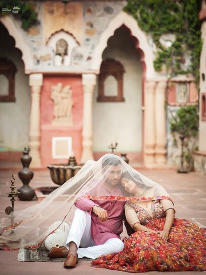 A moment of serene togetherness, draped under a delicate veil. This composition uses the heritage courtyard and traditional attire to tell a story of classic Indian romance.
