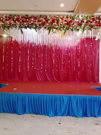 I can create beautiful stages for different budgets. This setup uses a simple frame decorated with a full floral arch, a bright red fabric backdrop, and hanging white tassels to create a festive look.