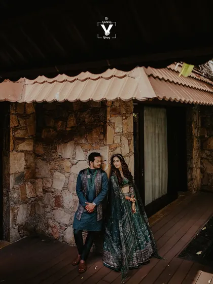A stylish portrait of the couple against a rustic stone and wood background. The groom's deep blue outfit and the bride's matching lehenga create a coordinated and fashionable look.