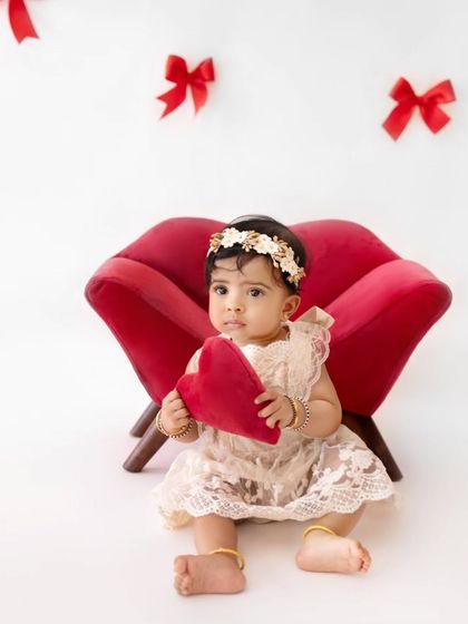 A perfect little valentine. This baby girl is ready to celebrate love, holding a heart-shaped pillow in a sweet and simple red-and-white themed setup.