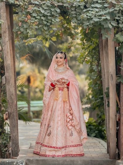 A beautiful full-length portrait of the bride, Simran, in her pastel pink lehenga, standing under a green archway, the picture of grace.