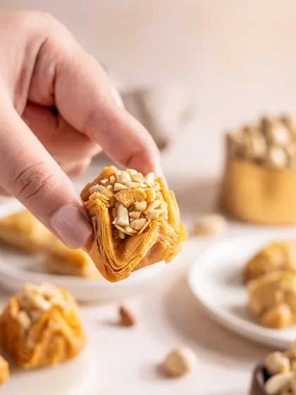 A hand holding up a piece of nutty baklava. This simple action brings the viewer into the scene and focuses on the delicious, crunchy texture of the dessert.