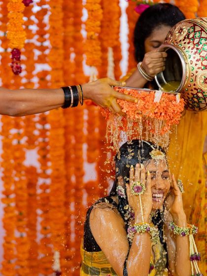 A moment of laughter and tradition during the Haldi ritual. The bride is showered with turmeric water against the backdrop of vibrant marigold strings, capturing the fun spirit of the event.