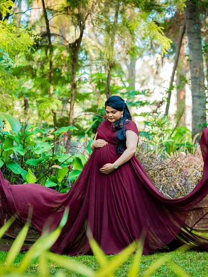 A serene solo maternity portrait with a flowing maroon gown in a lush green setting.