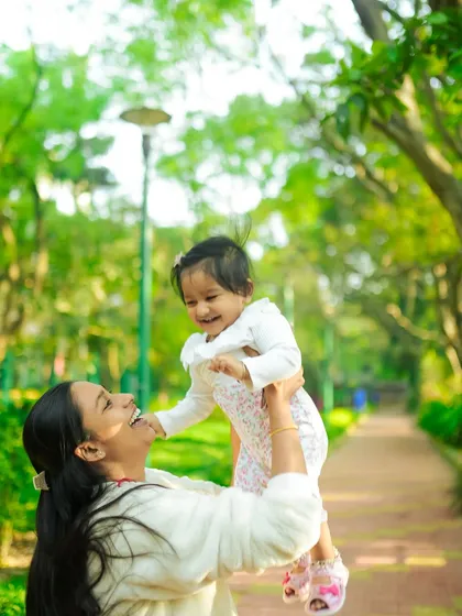 A mother lifting her daughter up in the air, both sharing a beautiful, happy moment. The lush green background of the park makes this a vibrant and joyful family portrait.