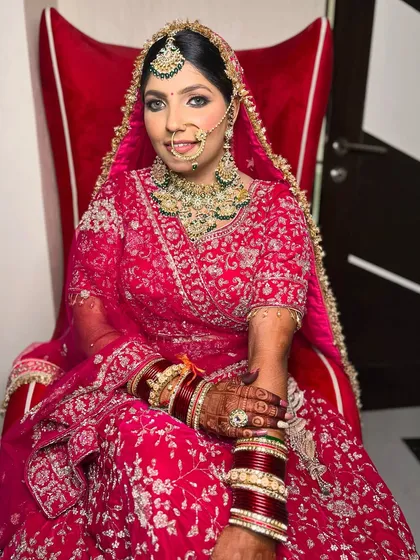 A beautiful portrait of a bride in a pink lehenga, seated in a grand red chair. Her makeup is soft and glowing.