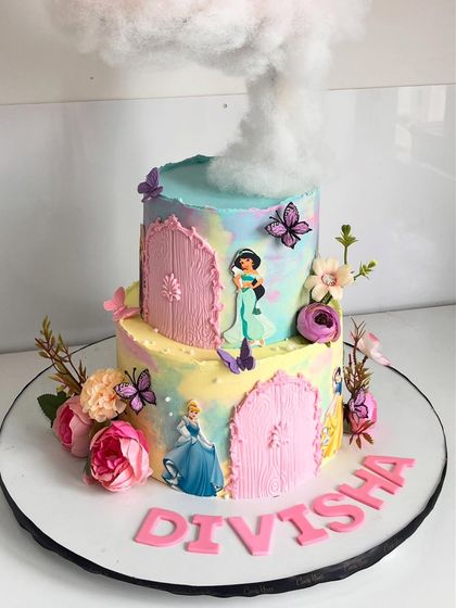The bottom tier of the princess castle cake, featuring beloved characters like Cinderella and Jasmine, set against a pastel watercolor background with floral and butterfly accents.
