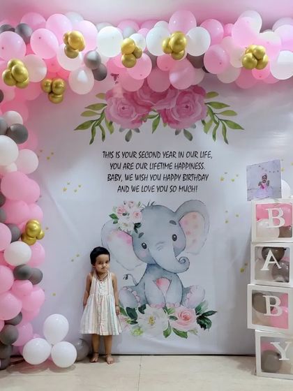 An adorable elephant-themed birthday party. The large, personalized backdrop is framed by a pink, white, and grey balloon arch, with "BABY" blocks on the side.