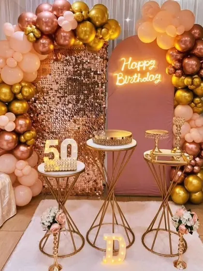 A glamorous 50th birthday setup featuring a rose gold sequin wall, a balloon arch in metallic colors, and elegant cake stands. The marquee numbers and neon sign complete the look.