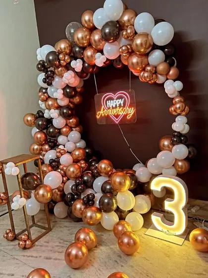 Celebrate your years together with a modern and stylish anniversary decoration. This circular balloon arch in a chic rose gold, white, and black theme, complete with a 'Happy Anniversary' neon sign and a light-up number, makes for a perfect photo backdrop.