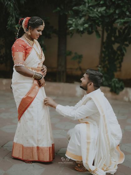 A tender moment as the groom kneels before his bride, adjusting her saree.