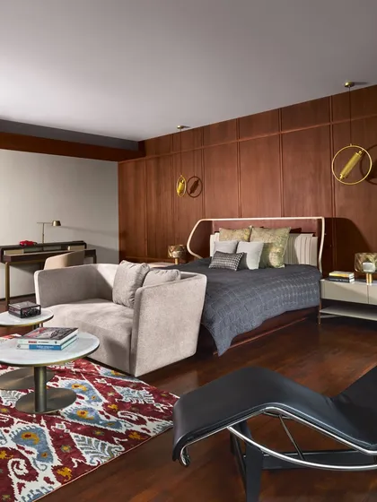 A bedroom at the Baid Residence featuring a rich wood-paneled wall and a Cassina chaise lounge. The design uses a warm, masculine color palette and a mix of modern and classic furniture.