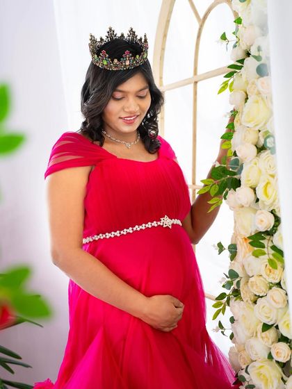 A quiet moment by the window. The mom-to-be in a vibrant pink gown looks down at her bump, framed by a wall of white roses, creating a soft and romantic mood.