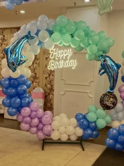 A circular balloon backdrop frame decorated with an ocean-colored garland and dolphin foil balloons. This modern setup, complete with a neon sign, is a popular choice for trendy birthday parties.