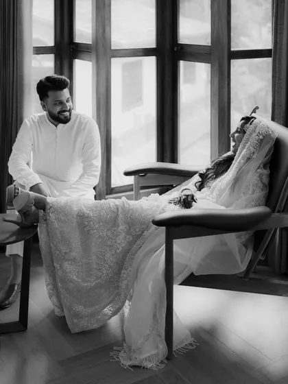 A candid, joyful moment shared between a bride and groom. The groom helps with her shoes, and their relaxed laughter in this black and white photo shows the comfortable, easy love they share.
