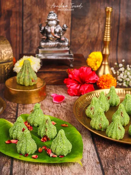 Green Pista Modaks arranged on a banana leaf. The rustic wooden background and scattered flowers give this shot a very natural and earthy vibe.