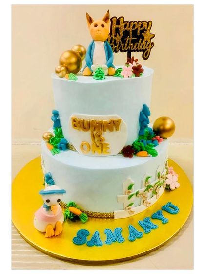 A sweet two-tiered cake for a first birthday with a "Bunny is One" theme. It features an adorable bunny topper, a little duck, and a charming white picket fence.