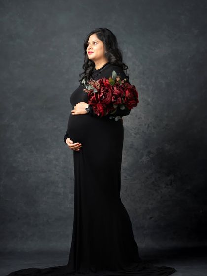 A powerful and classic maternity portrait. The mom-to-be stands tall and proud in a black gown, holding a bouquet of rich red flowers.