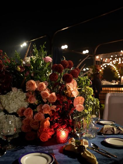 A close-up of a floral arrangement on a guest table, featuring a mix of red and peach roses against a dark blue tablecloth.