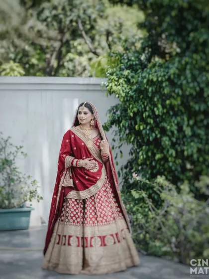 A full-length outdoor portrait of the bride in her traditional red and gold lehenga, looking elegant and poised.