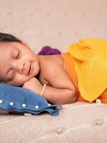 A simple and sweet pose on a soft blanket. The baby is sleeping on a small blue pillow, partially wrapped in colorful cloths, showcasing their tiny features and peaceful expression.