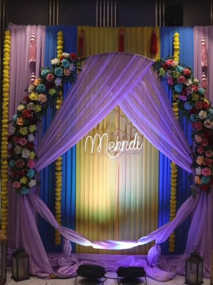 A modern Mehandi stage with a lavender theme. The setup includes a floral arch, a "Mehndi" neon sign, and draped fabric, creating a stylish and contemporary look.