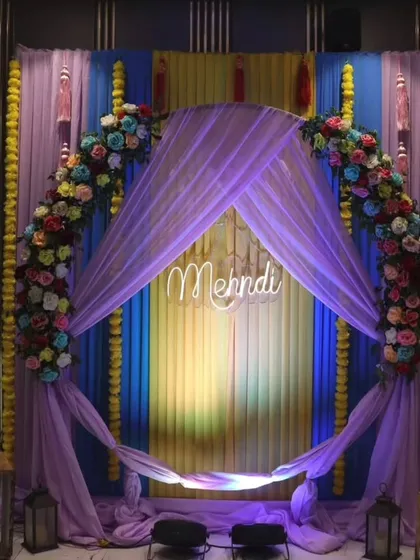 A modern Mehandi stage with a lavender theme. The setup includes a floral arch, a "Mehndi" neon sign, and draped fabric, creating a stylish and contemporary look.