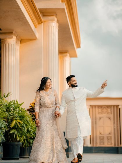 A couple walks together in a beautiful colonnade, dressed in elegant cream-colored outfits. The groom points towards their future, creating a hopeful and happy narrative.