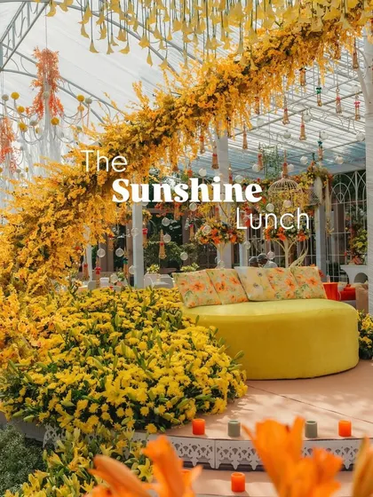 The main stage for 'The Sunshine Lunch', featuring a bright yellow curved sofa surrounded by a sea of yellow flowers. The setting is inside a greenhouse-like structure, bathing the entire scene in natural light.