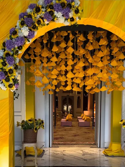 We love bringing vibrant traditions to life. This stunning Haldi ceremony entrance features a cascade of yellow fabric fans under an arch of bright sunflowers and purple blooms, welcoming guests into a joyous celebration.