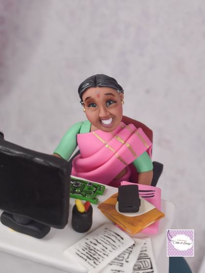 A close-up of a keepsake topper for a PCB engineer's 60th birthday. We replicated her work desk, including the computer, tiny circuit boards, and papers, with a smiling figurine of her in a beautiful pink saree.