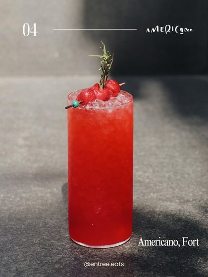 A bright, high contrast shot of a red cocktail against a simple concrete background. The direct sunlight makes the color pop, and the cherry and rosemary garnish adds a touch of freshness. It feels modern and refreshing.