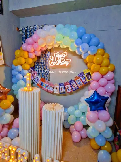 A pastel rainbow balloon arch for a first birthday, set up in a home. The decoration includes a photo banner, marquee number '1', and light-up letters for the name.