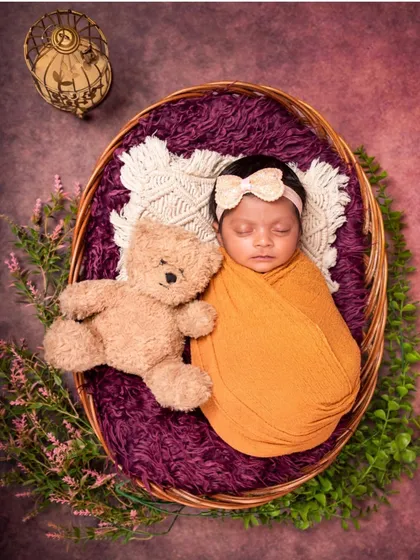 An overhead view of a newborn sleeping peacefully in a wicker basket. The combination of a mustard wrap, purple fur, and a teddy bear creates a colorful and sweet composition.